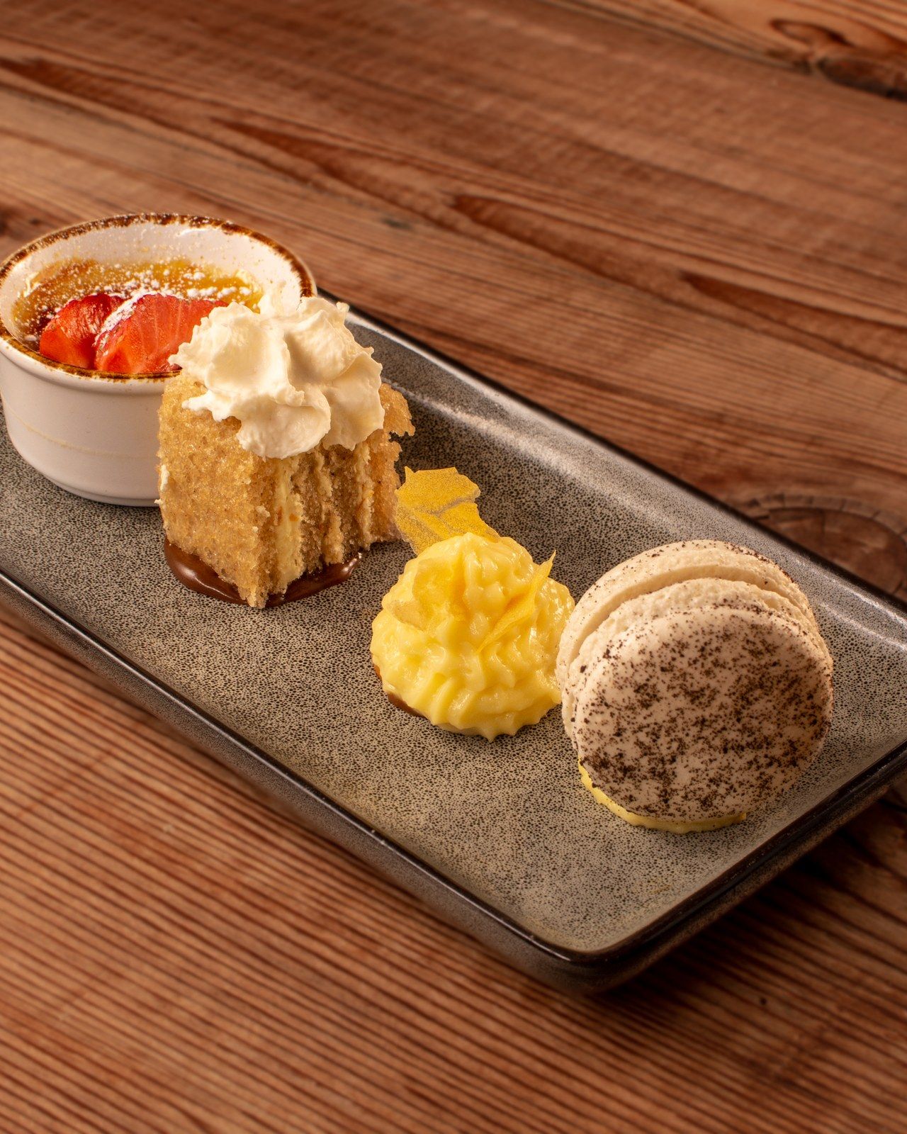 A dessert board of crème brûlée, cake with whipped cream, lemon curd and a macaron on a wood table